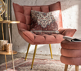 Een modern vormgegeven stoel met een fluweelachtige stof in een warme roze tint. De stoel heeft een gestoffeerde rugleuning en zitkussen, met extra kussens voor meer comfort. Een van de kussens heeft een uitgesproken oosters patroon met diverse kleuren. De stoel staat op elegante, gouden metalen poten die een chique contrast vormen met de zachte stof. De stoel staat op een Perzisch tapijt en naast een gouden bijzettafeltje met een ronde top. Op de achtergrond zie je een witte bakstenen muur die de industriële sfeer versterkt, terwijl de warme verlichting de ruimte een uitnodigend gevoel geeft. Rechts op de voorgrond zie je deels een gestapelde, ronde poef in een kleur die bij de stoel past.