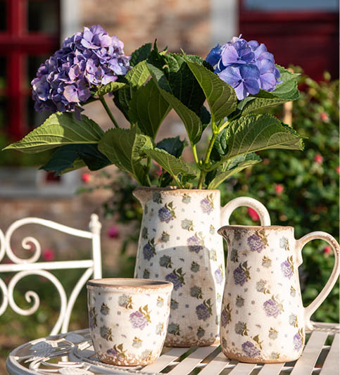 Een charmante set, bestaande uit twee kannen en een bijpassende bloempot, alle versierd met een delicaat bloemenpatroon in tinten van lila en groen, wat een landelijke uitstraling geeft. De items zijn tentoongesteld op een witte tuintafel met een gestreept patroon. Een volle bos hydrangea’s (hortensia's) in verschillende blauwtinten siert de kan en voegt een levendige toets van natuur toe. De setting is buiten, mogelijk op een veranda of in een tuin, met de schemerige achtergrond van een landhuis en een metalen tuinbank die een sfeer van rustieke elegantie creëren. Het is een idyllische weergave van buitenleven en ontspanning.